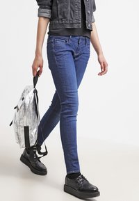 Jeans skinny azules combinados con una chaqueta corta negra, sosteniendo una mochila plateada. El calzado presenta detalles de malla negra y una suela de plataforma.