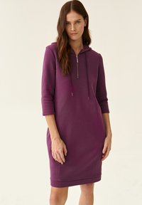 Kapucošované šaty v tmavej purpurovej, vyrobené z jemnej tkaniny. Má polovičný zips vpredu, rukávy po tri štvrtiny a bočné vrecká.