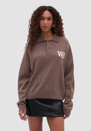 Mujer con cabello rizado que lleva un suéter marrón de manga larga con las letras "WB" y una minifalda de cuero negra, de pie frente a un fondo liso.