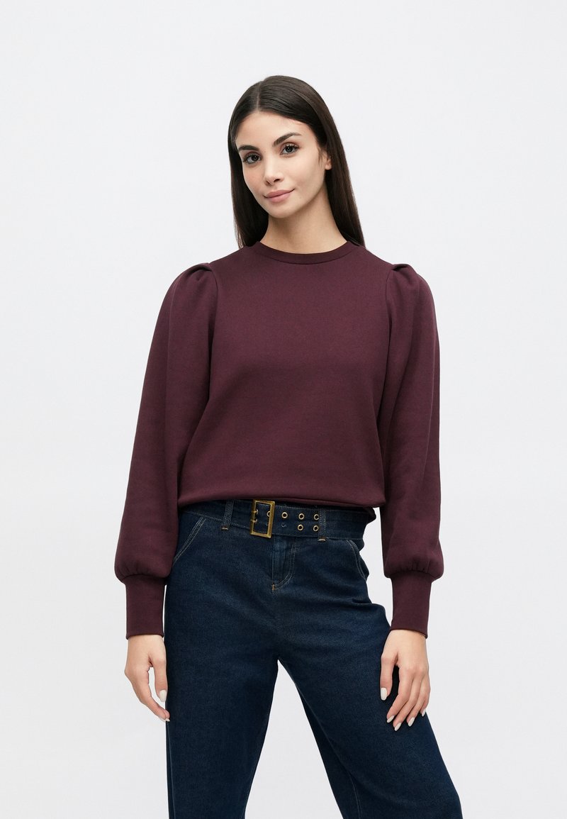 Mørkerød genser med puffede ermer; bomullsblanding, rund hals. Kombinert med mørkeblå høytlivs jeans med belte i midjen.