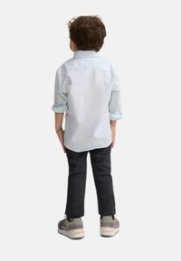 Camisa de cuadros de manga larga azul claro, con las mangas remangadas; pantalones oscuros, zapatillas gris con acentos multicolores; vista desde atrás.