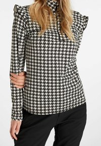 Donna che indossa una maglietta a righe bianche e nere con maniche lunghe e spalle arricciate, con zip a metà, abbinata a pantaloni neri, su uno sfondo bianco.