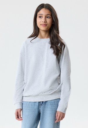 Pige med langt mørkt hår iført lys grå hættetrøje og lyseblå jeans, stående mod en ensfarvet hvid baggrund.