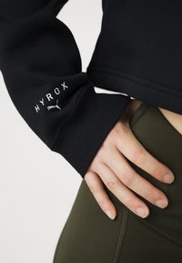 Felpa nera con polsini a coste, con logo "HYROX" ricamato in bianco, abbinata a leggings verde oliva. Inquadratura ravvicinata della mano sulla vita.