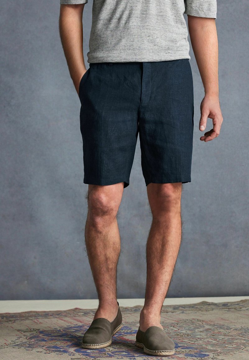 Marineblaue Leinenkurzshorts mit Seitentaschen, entspannter Schnitt und einem Saum, der knapp über dem Knie endet. Kombiniert mit einem grauen Kurzarmhemd.