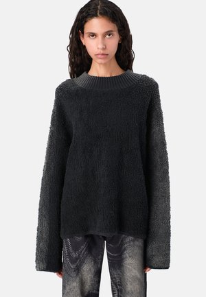 Mujer con cabello largo y rizado, vistiendo un suéter negro de textura oversized y pantalones con un patrón abstracto, de pie frente a un fondo blanco.