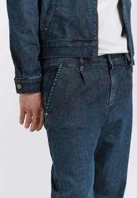Jeans in denim scuro abbinati a una giacca coordinata. Le caratteristiche includono cuciture gialle, dettagli con bottoni e tasche frontali. Tessuto liscio e morbido.