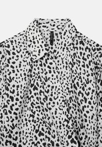 Camicia bianca con macchie leopardate nere irregolari, colletto a punta e etichetta nera visibile all'interno del colletto.