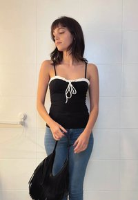 Čierny priliehavý top s bielymi volánmi a viazaním, v kombinácii s modrými džínsami. Drží čiernu kabelku s franžou. Pozadie s bielymi dlaždicami.