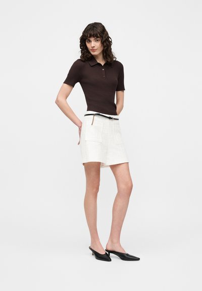 Femme portant un polo marron foncé ajusté à manches courtes, un short blanc taille haute avec poches et ceinture, et des talons noirs à bout pointu.