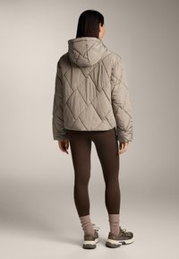 Giacca trapuntata grigia chiara con cappuccio, caratterizzata da un design ampio e da una cucitura diagonale evidente; indossata sopra leggins marroni e sneakers.