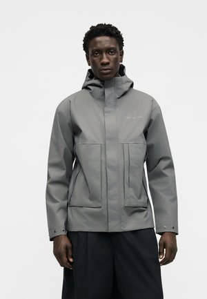 Homme portant une veste de pluie Columbia grise avec une capuche et deux grandes poches avant, debout devant un fond blanc uni.