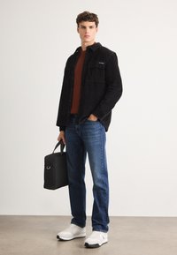 Camicia di velluto a coste nera, maglione di lana arancione, jeans di denim blu, sneakers bianche, che tiene una borsa nera. Vestibilità slim con dettagli a bottoni e tasche.
