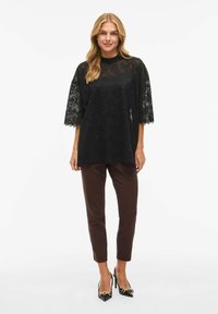 Czarna koronkowa bluzka z rozkloszowanymi rękawami, zestawiona z brązowymi obcisłymi spodniami i czarnymi szpilkami z noskami w szpic z dodatkiem złotych łańcuszków.