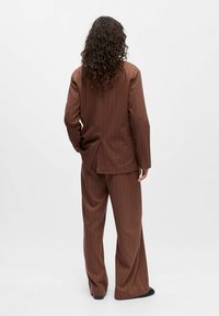 Personne avec de longs cheveux bouclés portant un costume en velours côtelé brun, de dos, se tenant devant un fond blanc uni.