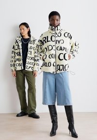 Veste en crème floue avec texte noir, fermeture éclair à l'avant, col montant ; le modèle porte un short en denim bleu à jambes larges et des bottes noires jusqu'au genou.
