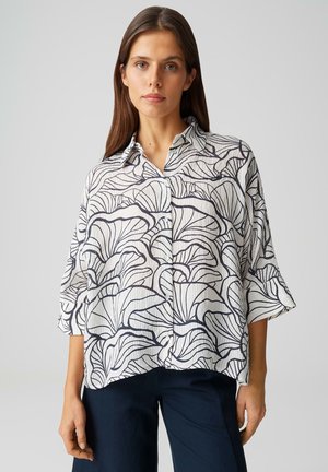 Femme aux longs cheveux bruns portant un chemisier blanc ample orné de lignes florales abstraites noires et un pantalon foncé, debout devant un fond uni.