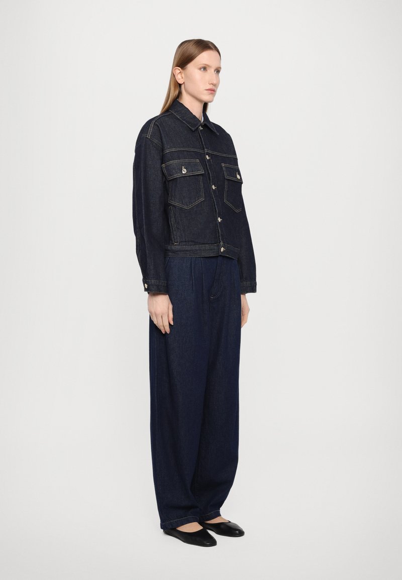 Giacca di jeans scura con due tasche anteriori, chiusura a bottoni e cuciture a vista, abbinata a pantaloni larghi in jeans scuri e scarpe flat.