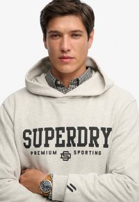 Sudadera con capucha gris claro con el logo "SUPERDRY" en negro, usada sobre una camisa de cuadros. Cuenta con un bolsillo delantero y puños acanalados. Reloj notable en la muñeca.