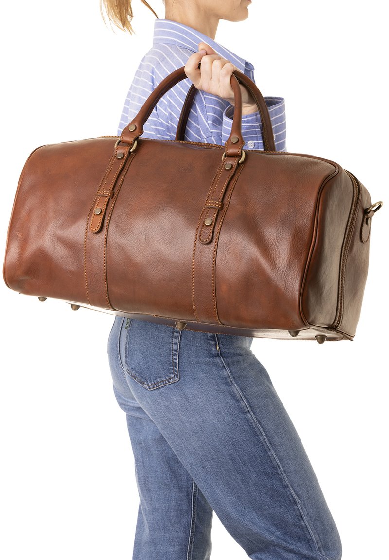 Sac de voyage en cuir marron avec doubles poignées, accessoires en métal, fermeture éclair sur le côté et détails de couture subtils ; texture lisse avec une forme structurée.