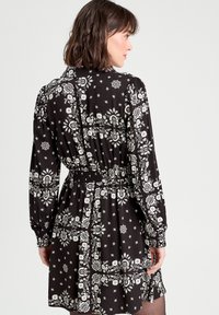 Robe noire avec des motifs floraux blancs, à manches longues, taille smockée et col de chemise. Présente une coupe décontractée et arrive au genou.