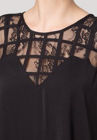 Schwarzes Top mit einem geometrischen Spitzenpanel und V-förmigem Ausschnitt, das eine Mischung aus festem Stoff und filigraner Spitzenstruktur hervorhebt.
