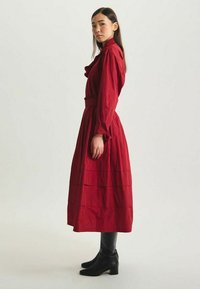 Robe midi rouge avec des manches longues et un col à volants. Présente une taille ceinturée et un design à niveaux. Associée avec des bottes noires jusqu'aux genoux.