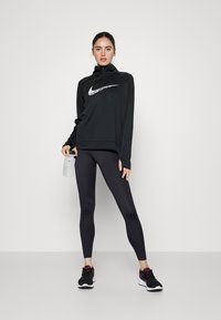Felpa nera da atletica con collo a zip, con logo Nike bianco. Leggings neri e scarpe da corsa nere con accenti rosa. Bottiglia d'acqua trasparente.