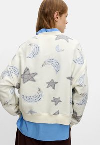Felpa color crema chiaro con un motivo giocoso di stelle e lune grigie, caratterizzata da una finitura testurizzata. Indossata sopra una camicia blu con colletto.