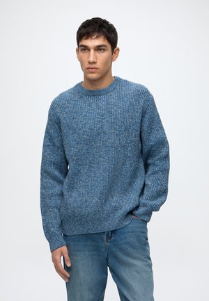 Jeune homme aux cheveux courts et foncés portant un pull en laine bleu et un jean bleu, debout avec une main dans la poche sur un fond clair.