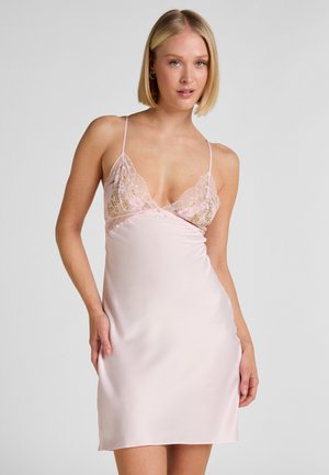 Vrouw draagt een lichtroze satijnen slipjurk met kanten rand en dunne bandjes, staand tegen een effen lichtgrijze achtergrond.