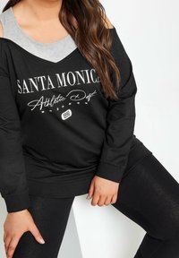 Czarna bluza z długim rękawem i odkrytymi ramionami, z białym napisem "Santa Monica Athletic Dept". W połączeniu z czarnymi legginsami.