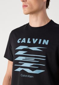 Juodas medvilninis marškinėlis su ryškiu mėlynu grafiniu spaudiniu "CALVIN" ir dinamiškomis linijomis, su apvalia iškirpte ir trumpomis rankovėmis.