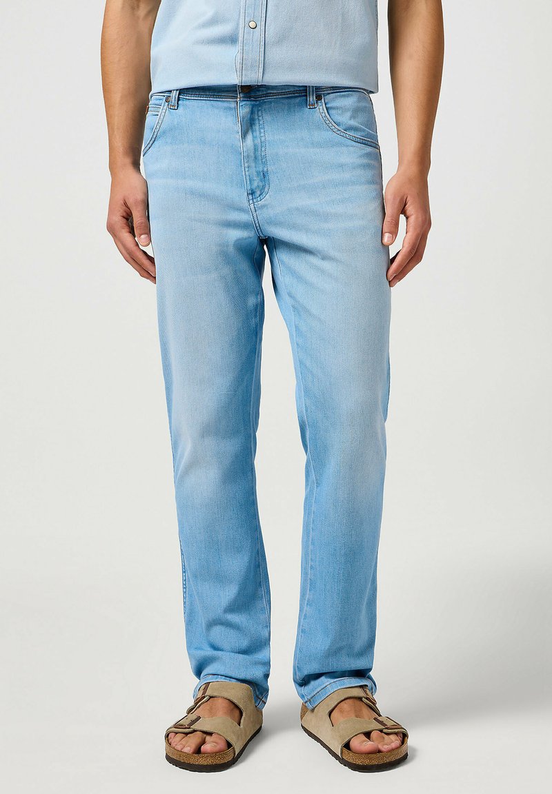 Jeans de denim azul claro con un corte recto, que presentan un diseño clásico de cinco bolsillos y un desvanecimiento mínimo. Llevados con sandalias marrones.