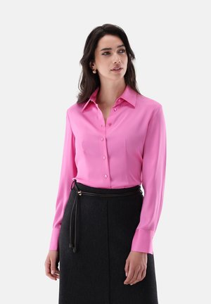 Donna che indossa una camicia rosa acceso con bottoni e una gonna nera con una sottile cintura nera, guarda leggermente di lato con un'espressione neutra.