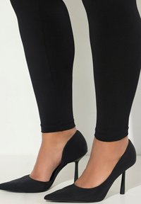 Scarpe nere con tacco a spillo, dalla punta appuntita e dalla finitura opaca, abbinate a leggings neri aderenti che mettono in mostra una silhouette slanciata.
