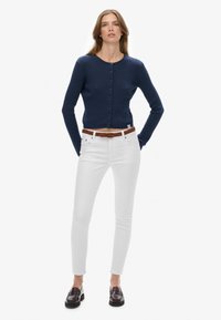 Kobieta stojąca z rękami za plecami, ubrana w granatowy zapinany kardigan, białe skinny jeansy z brązowym paskiem i ciemne loafersy.