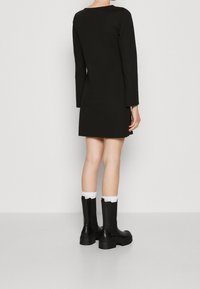 Svart långärmad klänning med rak skärning, parat med knähöga svarta stövlar och vita ankelsockor, visad bakifrån.