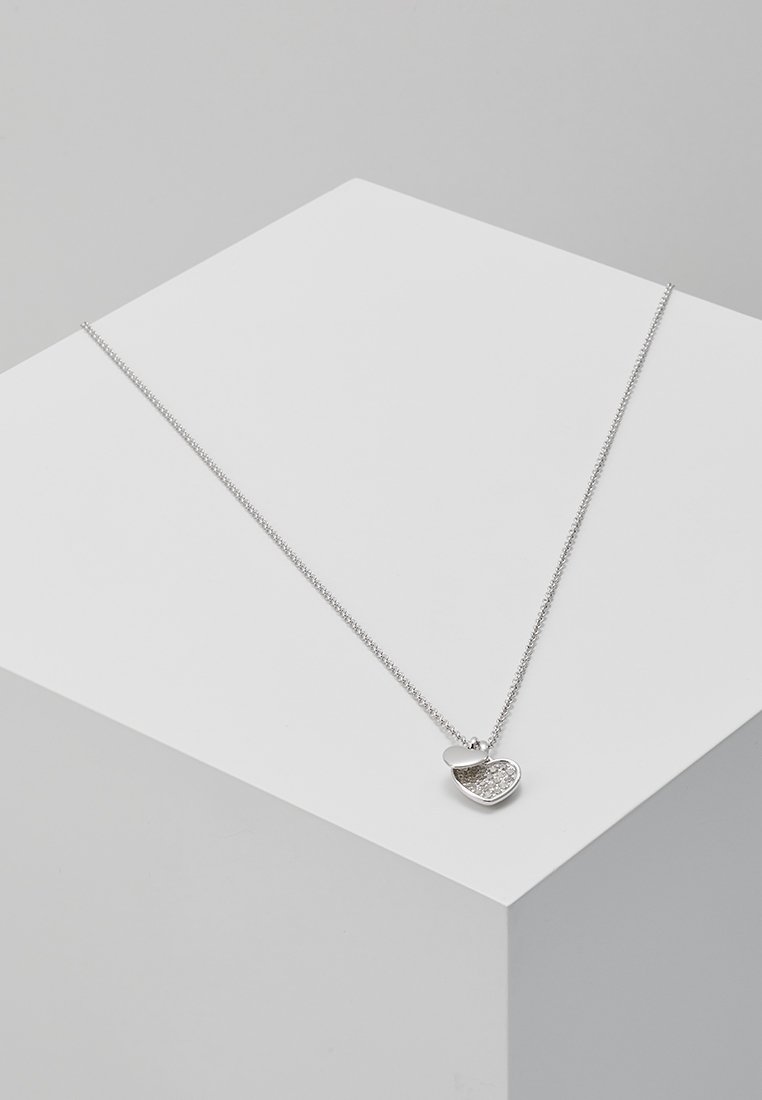 Collier délicat en argent avec deux pendentifs en forme de cœur, l'un uni et l'autre incrusté de petits cristaux, présenté sur un bloc géométrique blanc.