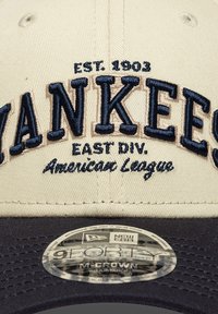Καπέλο μπέιζμπολ με κρεμ κορώνα και σκούρο ναυτικό γείσο. Κεντημένο κείμενο που περιλαμβάνει «YANKEES», «EST. 1903» και «East Div.» με αυτοκόλλητο λογότυπο MLB.