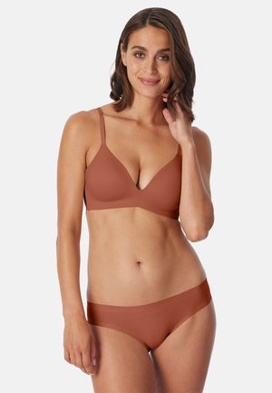 Frau mit schulterlangem braunem Haar, die einen nahtlosen BH in Rostfarbe und passende Unterwäsche trägt, lächelt mit der Hand nahe am Gesicht vor weißem Hintergrund.