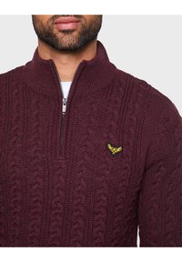 Burgunderfarbener gestrickter Pullover mit hohem Kragen, halbem Reißverschluss und gelbem Emblem auf der Brust, das ein strukturiertes Kabelstrickmuster aufweist.