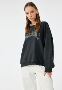 Czarny oversize sweter z ściągaczami u dołu i na rękawach. Posiada srebrny nadruk "Magical" w stylu pisma ręcznego. W zestawie z jasnymi spodniami.