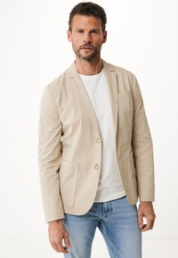 Beige Blazer-Jacke mit zwei vorderen Taschen, einreihigem Design und leichter Textur, kombiniert mit einem weißen T-Shirt und hellblauen Jeans.