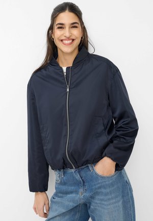 Lächelnde Frau mit langen dunklen Haaren trägt eine marineblau gestreifte Bomberjacke mit Reißverschluss und blaue Jeans, steht vor einem schlichten weißen Hintergrund.