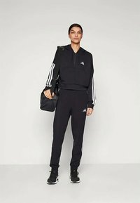 Žena v čiernom tréningovom obleku Adidas s bielymi pruhmi, drží čiernu kabelku cez rameno, stojí na svetlom pozadí.