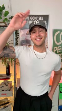 Valkoinen istuva t-paita, hopeaketju ja mustat housut; päällään harmaa lippalakki logolla, hymyilee ja wave. Sisätilat kasvien kanssa.