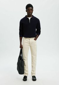 Marineblauer Strickpullover mit Reißverschlusskragen, cremefarbene Hose, schwarze Schuhe und dunkelgraue Tragetasche. Fokus auf Textur und Farbkontrast.