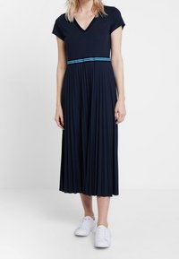 Marinblå, plisserad klänning med V-urringning, korta ärmar och ett kontrasterande blått horisontellt rand vid midjan. Kombinerad med vita sneakers.