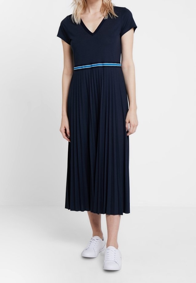 Marinblå, plisserad klänning med V-urringning, korta ärmar och ett kontrasterande blått horisontellt rand vid midjan. Kombinerad med vita sneakers.